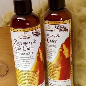 May include: Two brown glass bottles of rosemary and apple cider hair spray. The bottles have a white label with a red illustration of a woman with long hair. The label reads "Rosemary & Apple Cider Spray for Hair" and lists ingredients. The bottles are 4.5 fl oz.
