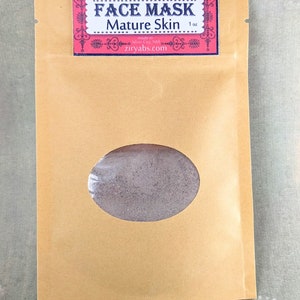 May include: A brown paper pouch with a clear window showing a gray powder inside. The pouch has a red and white label that reads "FACE MASK Mature Skin 1 oz made in Silver City, NM ziryabs.com"