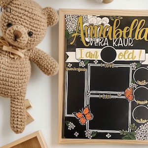 May include: A black chalkboard with a floral design and gold accents. The text "Annabella Vera Kaur I am old!" is written on the board. There are sections for favourite foods, things I love, things I can do, teeth, pounds, and inches.