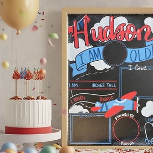 May include: A birthday-themed chalkboard sign with the name "Hudson" in red, and the words "I AM OLD!" in blue. The sign includes sections for height in inches, weight in pounds, and favorite foods. A white cake with red accents and decorative toppers is in the foreground.