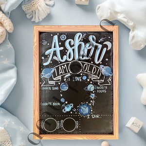 May include: A wooden framed chalkboard with a space-themed design. The chalkboard says "Asher I am [age] old" and has sections for favorite song, book, foods, and what the child can do. The chalkboard is on a light blue background with a white star and a stuffed animal.