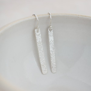May include: A pair of silver dangle earrings with a floral pattern. The earrings are long and rectangular in shape.
