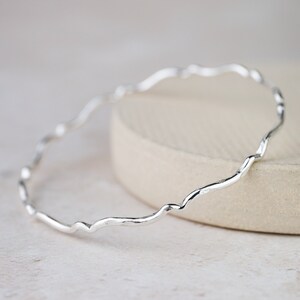 May include: A silver bangle bracelet with a wavy, organic design. The bracelet is made of a thin, delicate metal and has a smooth, polished finish.