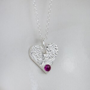 May include: A silver necklace with a heart-shaped pendant featuring a detailed floral design and a pink gemstone in the centre.