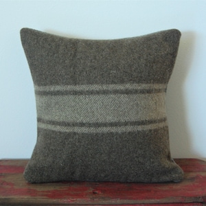 May include: Square pillow in shades of brown and tan. The pillow has a textured, woven appearance with a horizontal stripe design across the center. The pillow is resting on a red, weathered wooden surface.