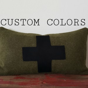 May include: Olive green rectangular pillow with a large black cross design. The pillow is displayed on a distressed red surface. Text above the pillow reads "CUSTOM COLORS".