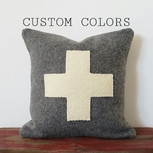 May include: Gray wool pillow with a white felt cross in the center. "CUSTOM COLORS" is written in black text at the top of the image.