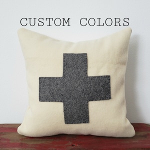 May include: Cream-colored decorative pillow with a large gray cross design. The text "CUSTOM COLORS" is above the pillow. The pillow is on a red surface.