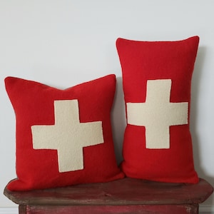 May include: Two red throw pillows with a cream-colored cross design. One pillow is square, and the other is rectangular. The pillows are placed on a distressed red wooden surface. The pillows are a decorative accent.
