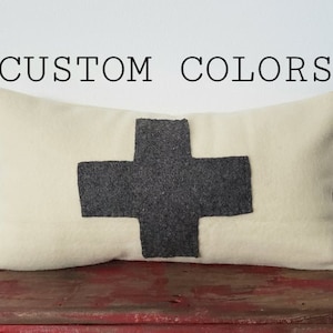 May include: Cream-colored rectangular pillow with a large, dark gray cross design. The text "CUSTOM COLORS" is printed in black above the pillow. The pillow is made of a soft, textured fabric.