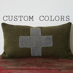 May include: Olive green rectangular pillow with a gray cross applique. The pillow is on a red wooden surface. The text "CUSTOM COLORS" is above the pillow. The pillow is a decorative home accent.