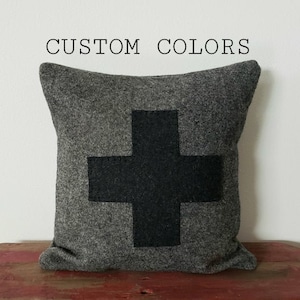 May include: Gray decorative pillow with a large black cross design. The pillow is made of a textured fabric and features the text "CUSTOM COLORS" at the top. The pillow is displayed on a red surface.