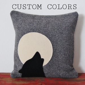 May include: Gray decorative pillow with a felt design of a howling wolf in front of a full moon. The moon is a cream color, and the wolf is black. The text "CUSTOM COLORS" is at the top.