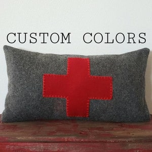 May include: A rectangular gray pillow with a large red cross stitched onto the front. The text "CUSTOM COLORS" is above the pillow. The pillow is resting on a red wooden surface.