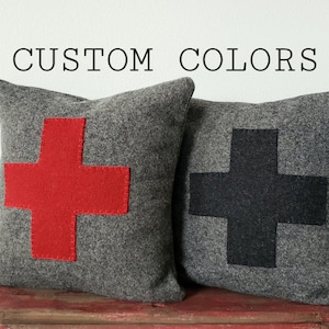 May include: Two gray throw pillows with large cross designs. One pillow has a red cross, the other a black cross. The pillows are on a red surface. The text "CUSTOM COLORS" is above the pillows.