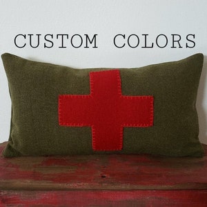 May include: Olive green rectangular pillow with a large red cross applique. The pillow is displayed on a distressed red wooden surface. The text "CUSTOM COLORS" is above the pillow.