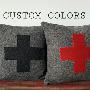 May include: Two gray throw pillows with large cross designs. One pillow has a black cross, the other a red cross. The pillows are on a rustic red surface against a white background. Text above reads "CUSTOM COLORS".