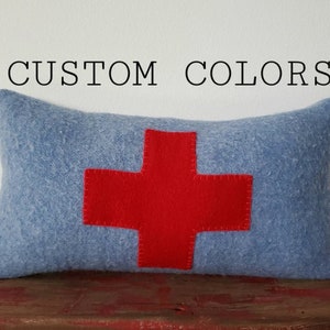 May include: A rectangular, light blue pillow with a large red cross in the center. The text "CUSTOM COLORS" is above the pillow. The pillow is on a red wooden surface.