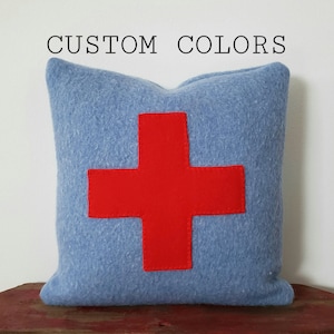May include: A light blue decorative pillow with a large red cross design. The pillow is made of a soft, textured fabric. The words "CUSTOM COLORS" are printed above the pillow.