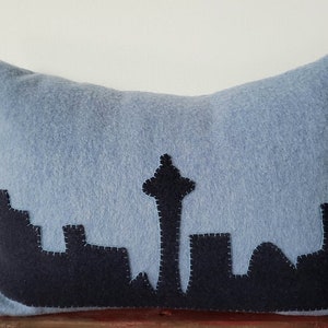 May include: A blue throw pillow with a black felt applique of the Seattle skyline, including the Space Needle.