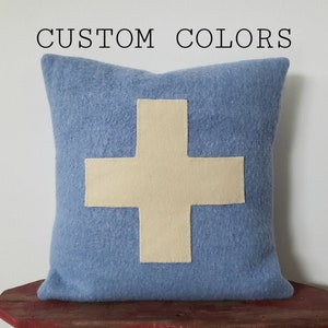 May include: A square, light blue pillow with a large, cream-colored cross design. The text "CUSTOM COLORS" is above the pillow. The pillow is made of a soft, textured material and is likely for home decor.