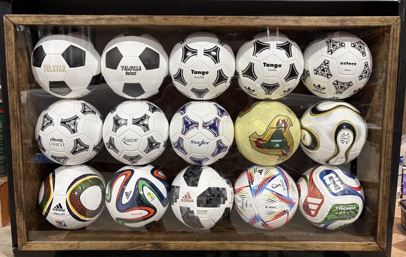 May include: A collection of soccer balls displayed in a wooden framed case. The balls feature various designs, including black and white panels, geometric patterns, and brand logos. Some balls have gold and colorful accents.
