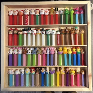 May include: A wooden display case filled with a collection of PEZ dispensers. The dispensers feature various characters, including Santa Claus, snowmen, bunnies, ducks, and Disney princesses. The dispensers are arranged on multiple shelves with red, green, and other colored stems.
