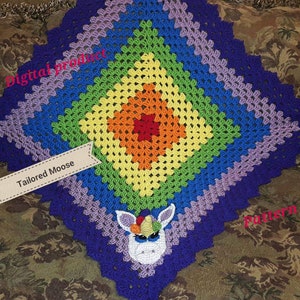 Op de afbeelding: Een regenboog gehaakte deken met een eenhoorn ontwerp in het midden. De deken is gemaakt met verschillende gekleurde garen, waaronder paars, blauw, groen, geel, oranje en rood. De eenhoorn is wit met een roze hoorn en een blauwe manen. De tekst "Tailored Moose" staat op een wit label.