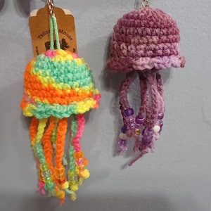 May include: Two handmade crochet jellyfish keychains. One is multicolored with orange, green, and yellow. The other is purple with beaded tentacles. The keychains are hanging from a metal ring.