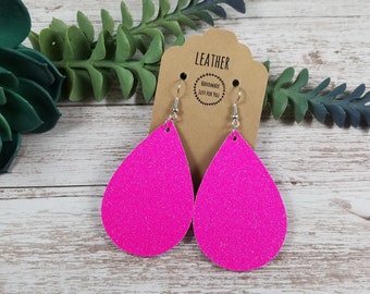 May include: A pair of pink glitter teardrop shaped leather earrings with silver hooks. The earrings are on a brown card with the text "LEATHER HANDMADE JUST FOR YOU".