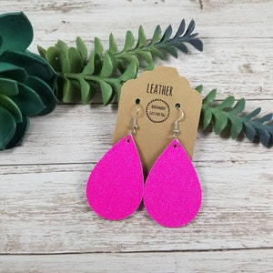 May include: A pair of pink glitter teardrop earrings with silver hooks. The earrings are on a brown card with the text "LEATHER HANDMADE JUST FOR YOU".