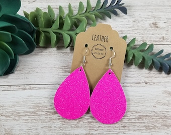 May include: A pair of pink glitter teardrop earrings with silver hooks. The earrings are on a brown card with the text "LEATHER HANDMADE JUST FOR YOU".