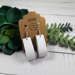 Puede incluir: Pendientes de cuero blanco con ganchos plateados. Los pendientes están en una tarjeta marrón con el texto "LEATHER HANDMADE JUST FOR YOU".