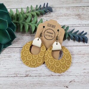 Pode incluir: Um par de brincos de couro amarelo mostarda com um padrão de favo de mel e detalhes em couro branco. Os brincos estão pendurados em um cartão marrom com o texto "Leather Handmade Just For You".