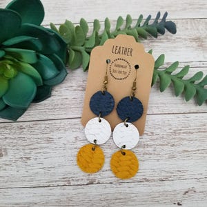 May include: Dangle earrings featuring three stacked circular leather discs in navy blue, white, and mustard yellow. The discs have a textured, braided-style pattern. The earrings are attached to bronze-toned metal hooks on a brown card labeled LEATHER.