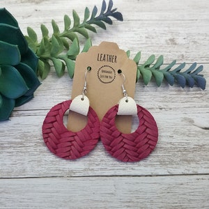 May include: A pair of red leather earrings with a woven design and white leather accents. The earrings are hanging from silver hooks. The earrings are on a brown card with the text "Leather Handmade Just For You".