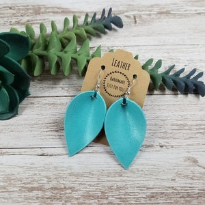 May include: Teal leather earrings with silver hooks. The earrings are shaped like a teardrop and have a smooth, matte finish. The earrings are handmade and come with a tag that says "Leather Handmade Just For You".