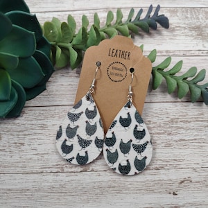 May include: A pair of teardrop-shaped leather earrings with a black and white chicken print. The earrings are hanging from silver hooks and have a brown paper tag that says "Leather Handmade Just for You".