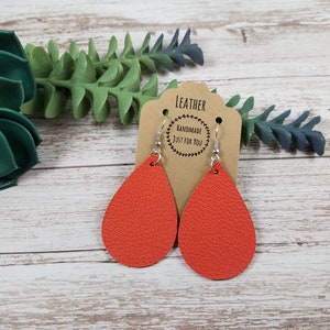 May include: A pair of orange leather teardrop earrings with silver hooks. The earrings are handmade and have a textured surface. The tag says "Leather Handmade Just For You".