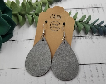May include: A pair of gray leather teardrop earrings with silver hooks. The earrings are on a brown card with the text "Leather Handmade Just for You".
