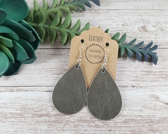 May include: A pair of gray leather teardrop earrings with silver hooks. The earrings are handmade and have a textured surface. The earrings are displayed on a brown card with the text "Leather Handmade Just for You".