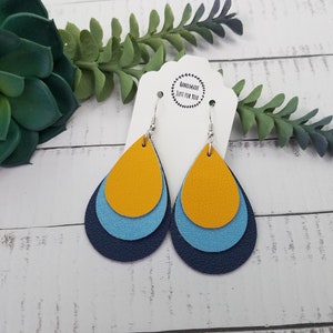 May include: A pair of teardrop-shaped earrings made of layered leather. The earrings are in shades of navy blue, light blue, and mustard yellow. The earrings are hanging from silver hooks.