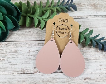 May include: A pair of blush pink teardrop earrings with silver-colored hooks. The earrings are displayed on a brown card with the word "LEATHER" and the text "HANDMADE JUST FOR YOU" printed on it. The background includes green and blue-green foliage.