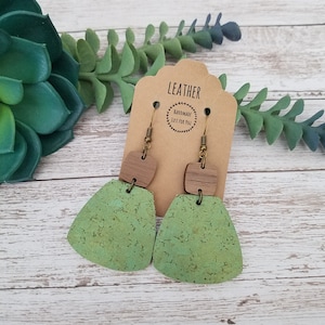 May include: Green and brown cork earrings with a geometric shape. The earrings are handmade and feature a small wooden square at the top. The earrings are hanging from a brown card with the text "LEATHER HANDMADE JUST FOR YOU".