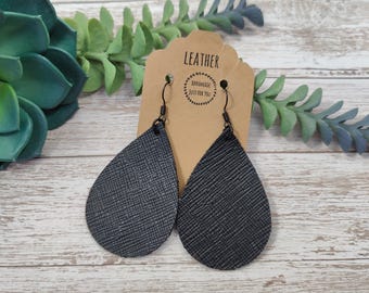 May include: A pair of black leather teardrop earrings with a textured finish. The earrings are hanging on a brown card with the word "LEATHER" and the text "Handmade Just For You".