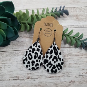 May include: A pair of black and white leopard print leather teardrop earrings with black hooks. The earrings are on a brown card with the text "LEATHER HANDMADE JUST FOR YOU".