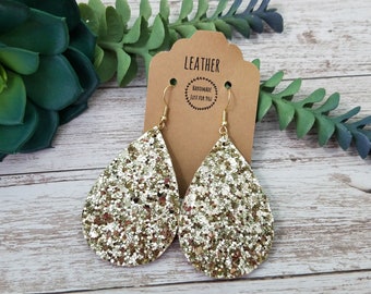 May include: Gold glitter teardrop earrings with gold hooks. The earrings are handmade and have a "Leather Handmade Just For You" tag.