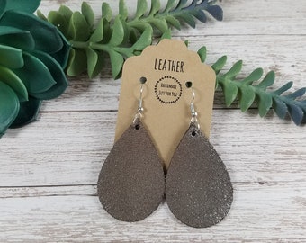 May include: A pair of silver teardrop-shaped leather earrings with a sparkly finish. The earrings are hanging from a brown card with the text "LEATHER HANDMADE JUST FOR YOU".