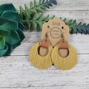 May include: A pair of mustard yellow leather earrings with a brown leather accent. The earrings are round and have a woven texture. The earrings are hanging from a brown card with the text "Leather Handmade Just For You".