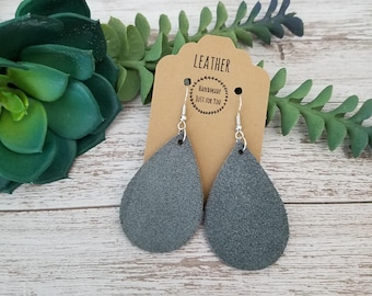 May include: Grey teardrop leather earrings with silver hooks. The earrings are displayed on a brown tag that reads "LEATHER" and "HANDMADE JUST FOR YOU". The background includes green plants and a light-coloured wooden surface.